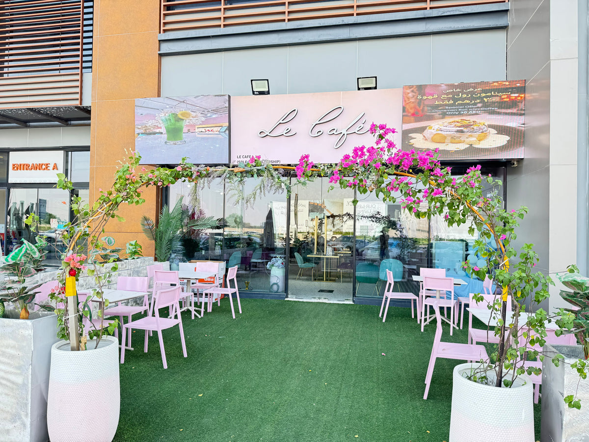 Outdoor cafe seating area with pink chairs and tables arranged on green artificial turf in front of a modern building. A pink sign reads "Le Cafe" above glass doors, with pink flowers arching overhead and large white planters with leafy plants at the sides.