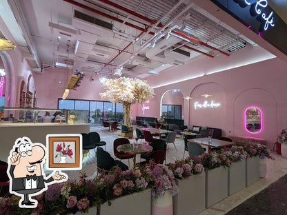 A pink-themed cafe interior with pink walls, neon lighting, arched windows, and multiple dining tables with chairs. A large floral tree centerpiece stands in the middle, and flower arrangements line the foreground.