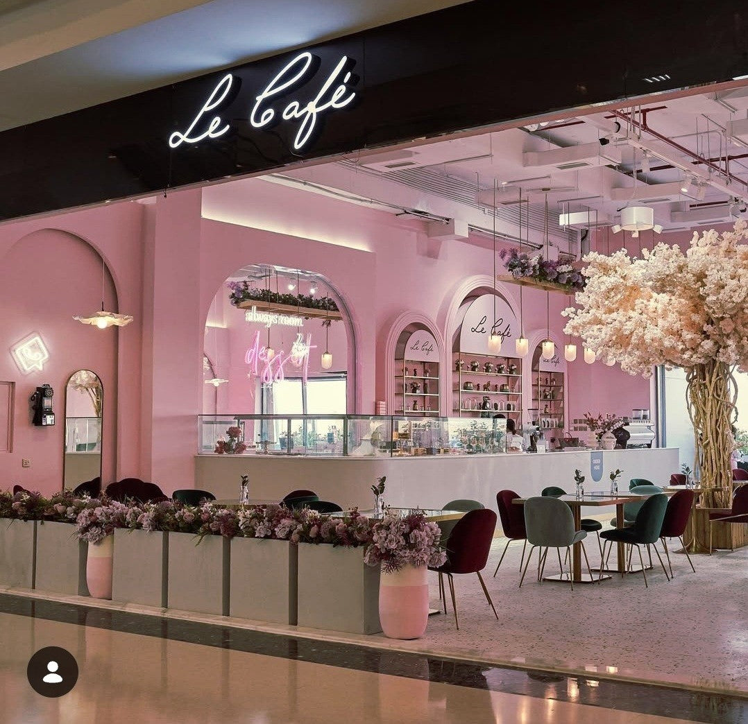 A pink cafe interior with arched windows, a long counter, neon sign, shelves, tables, chairs, floral arrangements, and a large flowering tree on the right. A black storefront sign at the top reads “Le café” in white script.