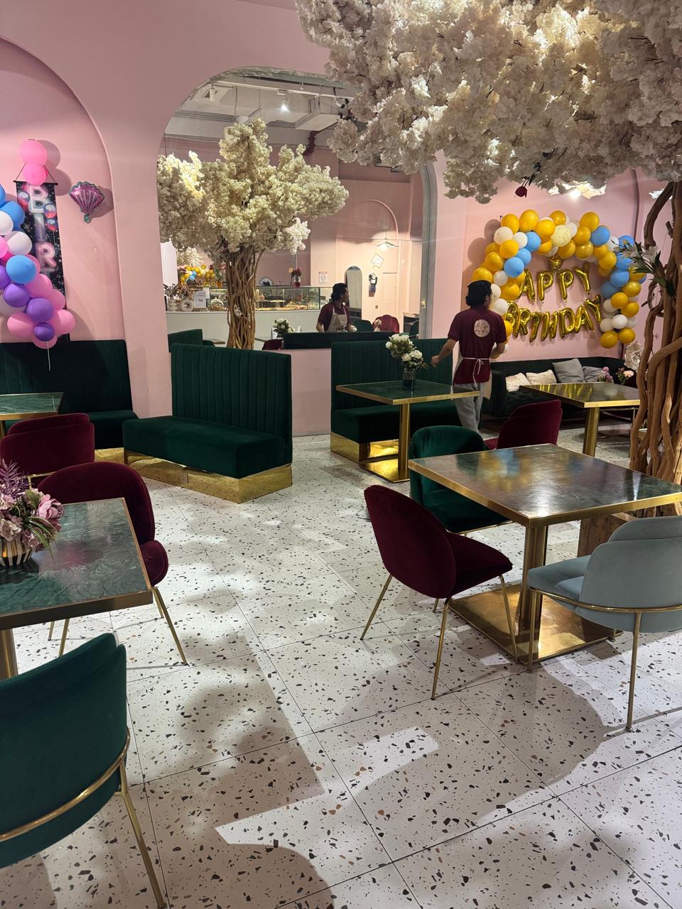 A decorated indoor event space or party venue with pink walls, white floral tree decorations, teal velvet booths, and multiple dining tables with burgundy, gray, and gold chairs. Balloons and a gold 'Happy Birthday' balloon garland are visible in the background.