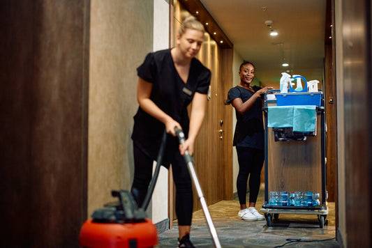 Two women cleaning a hallway with a vacuum cleaner and cleaning cart.